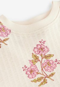 Haut couleur crème avec texture côtelée, orné de fleurs brodées roses et de feuilles vertes le long de l'encolure.