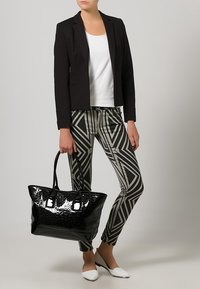 Kobieta w czarnym żakiecie, białej bluzce, spódnicach w geometryczny wzór, białych butach, trzymająca dużą, błyszczącą czarną torbę na dwa uchwyty.