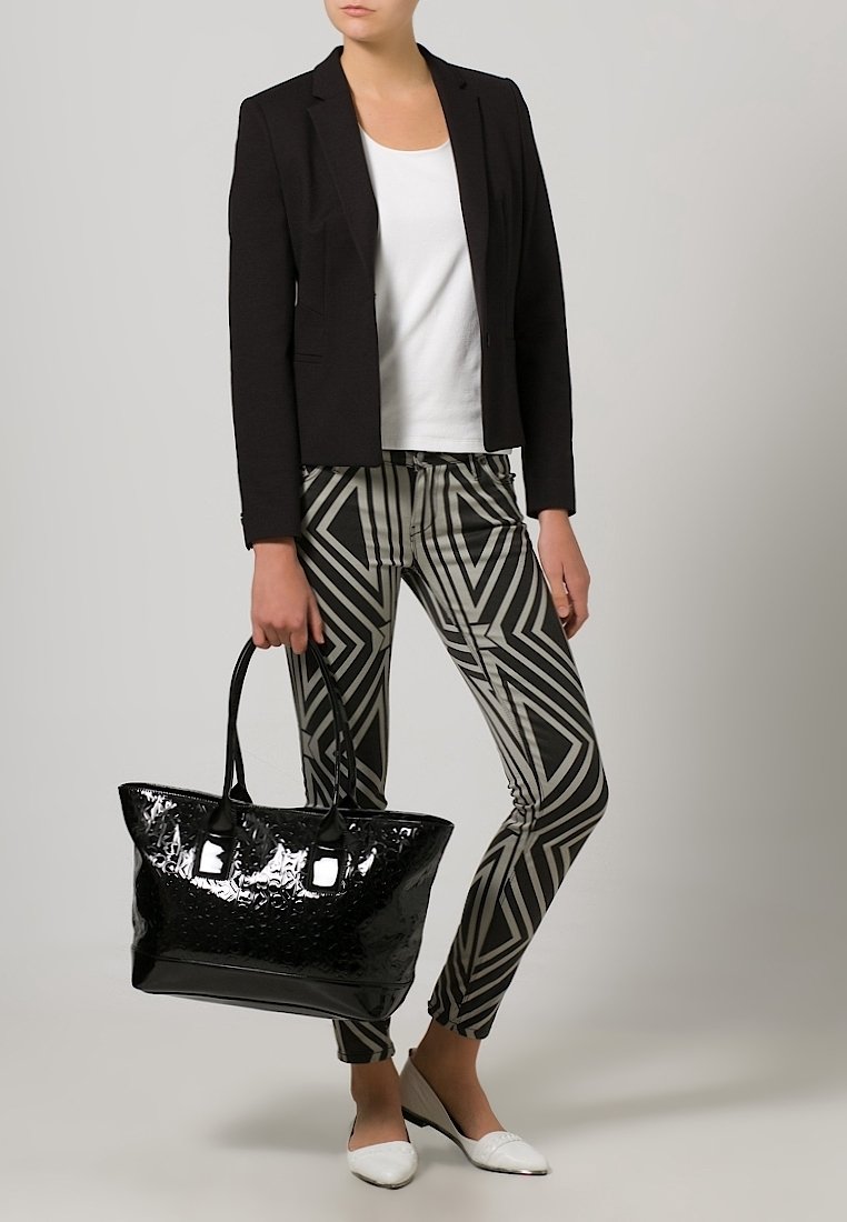 Kobieta w czarnym żakiecie, białej bluzce, spódnicach w geometryczny wzór, białych butach, trzymająca dużą, błyszczącą czarną torbę na dwa uchwyty.