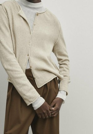 Beige knitted cardigan with a V-neck, five button closures, and ribbed cuffs, worn over a light gray turtleneck and paired with brown trousers.