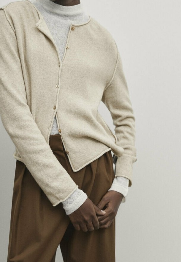 Cardigan beige a maglia con scollatura a V, chiusura a cinque bottoni e polsini a coste, indossato sopra un dolcevita grigio chiaro e abbinato a pantaloni marroni.