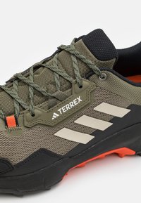 Chaussures de randonnée Terrex en vert olive avec des accents noirs. Dotées d'un dessus en maille texturée, d'œillets renforcés et d'une semelle extérieure orange contrastante.