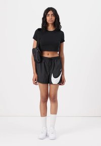 Melnā īsā T-kreklā, melnā sporta šortos ar baltu Nike logo, baltas zeķes un baltie apavi. Mazs melns plecu soma.