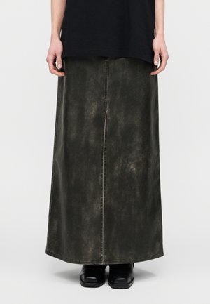 Jupe longue de couleur vert foncé, confectionnée en denim avec une texture délavée. Présente une couture frontale et une coupe droite. Portée avec des bottes noires.