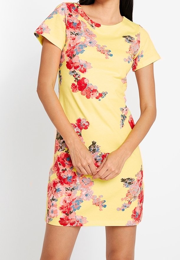 Femme portant une robe courte jaune avec des motifs floraux rouges, roses et bleus, debout, les mains jointes devant elle.