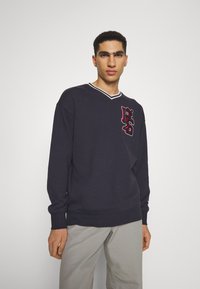 Jūras spēku garās piedurknes sporta krekls ar V-veida kakla izgriezumu, ar kontrastējošu baltu apdari un sarkanu un melnu izšūtu logotipu uz krūtīm. Mīksta tekstūra, brīva piegriezuma.