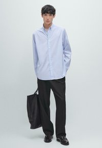 Camicetta a righe blu chiaro con colletto, abbinata a pantaloni larghi neri e scarpe in pelle nere. Il modello tiene in mano una borsa tote nera.