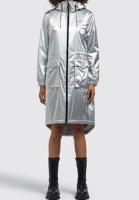 Srebrny wodoodporny płaszcz przeciwdeszczowy z kapturem, elastycznymi mankietami, dwiema przednimi kieszeniami i czarnym zamkiem błyskawicznym. Posiada dłuższy tylny brzeg.