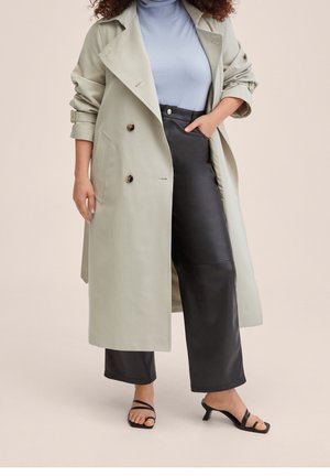 Femme portant un trench-coat gris clair, un col roulé bleu clair, un pantalon en cuir noir et des sandales à talons noires, debout avec une main dans la poche.