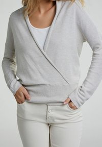 Un pull en maille léger gris clair avec un design croisé, des manches longues et une taille côtelée. Porté sur un haut blanc et associé à un pantalon crème.