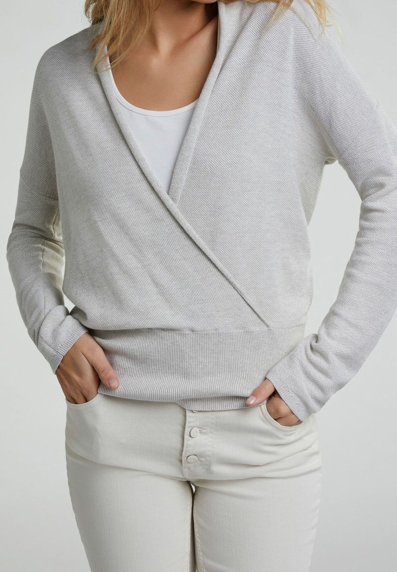 Un pull en maille léger gris clair avec un design croisé, des manches longues et une taille côtelée. Porté sur un haut blanc et associé à un pantalon crème.