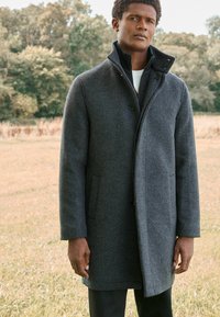 Abrigo de lana gris con cuello alto, cierre de botones y bolsillos laterales, que presenta una textura suave y un diseño a medida.
