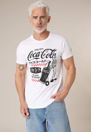 Homme d'âge moyen aux cheveux gris portant un t-shirt blanc avec un design vintage Coca-Cola et un jean bleu clair, debout devant un fond beige.