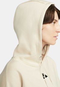 Felpa beige con cappuccio e cordini, decorata con un logo triangolare. Tessuto morbido e design aderente, dotata di un ampio cappuccio e polsini a costine.
