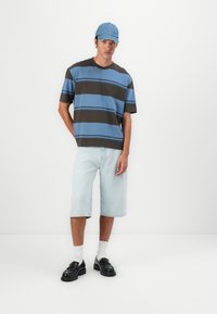 Striped t-shirt in blue and brown, light denim shorts, black loafers, white socks, blue cap. Casual outfit with relaxed fit design.