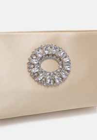 Satinbeige clutch med en cirkulär rhinestone-dekoration som har olika storlekar av klara stenar i silverfärgad metall.