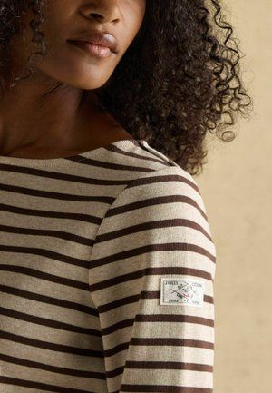 Striped long-sleeve top in beige and brown, featuring a round neckline and a fabric patch with a logo on the left sleeve. Soft texture.