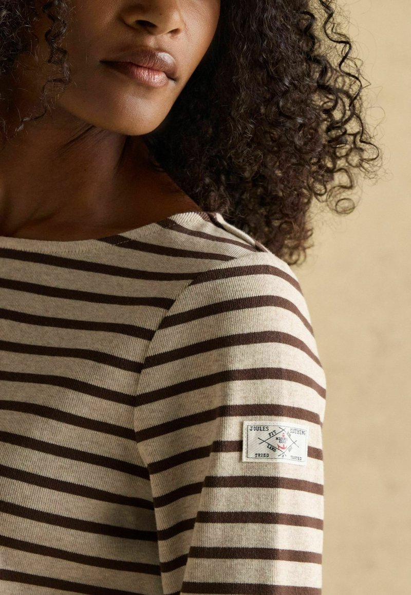 Gestreift Langarmshirt in Beige und Braun, mit Rundhalsausschnitt und einem Stoffaufnäher mit Logo am linken Ärmel. Weicher Stoff.