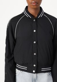 Chaqueta bomber negra con una textura ligera, presenta un cierre de botones en la parte frontal, cuello a rayas y cintura de canalé.