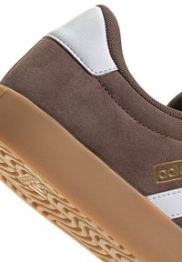 Sneaker in suede marrone con accenti in pelle bianca, logo dorato, suola in gomma testurizzata e dettagli lisci sul tallone e sui fianchi.