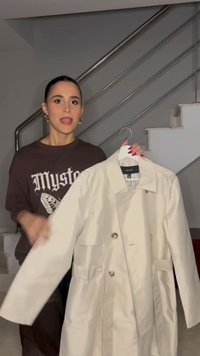 Žena s tmavými vlasy drží a předvádí béžový trenčkot na ramínku v schodišti s kovovým zábradlím.