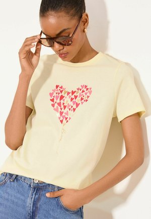 Kvinde justerer solbriller og har en lysegul t-shirt på med en hjerteformet samling af røde og pink hjerter og gyldent "love"-tekst, blå jeans.
