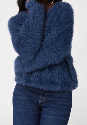 Femme portant un pull tricoté bleu duveteux et un jean en denim foncé, avec la main droite touchant son cou et des boucles d'oreilles en or visibles.