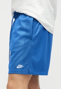 Μπλε αθλητικό σορτς Nike με ελαστική μέση και πλευρική τσέπη, φορεμένο από άτομο με λευκό πουκάμισο, εμφανίζεται από το μέσο του μηρού μέχρι τη μέση.