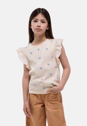 Jeune fille aux longs cheveux bruns portant un haut crème à manches volantées avec de petites fleurs bleues et un pantalon marron clair, main dans la poche.