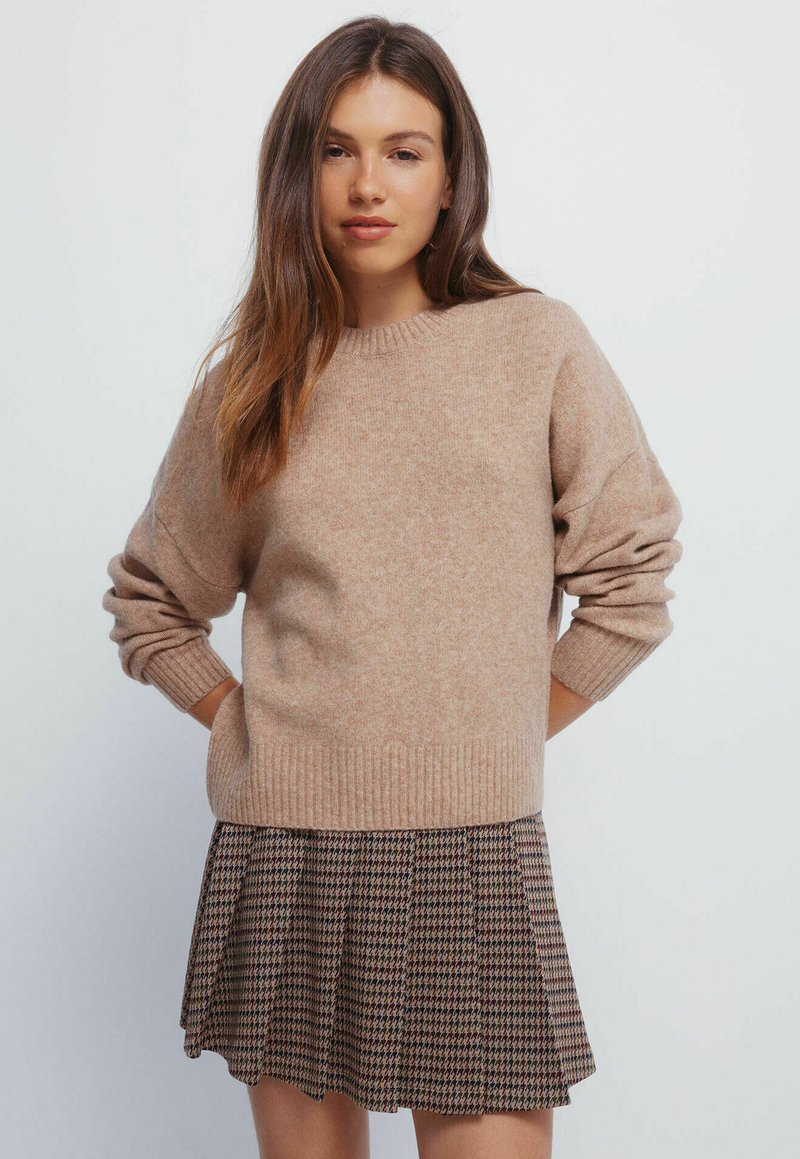 Maglione beige lavorato a maglia con polsini e orlo a coste, abbinato a una gonna plissettata a fantasia in toni scuri e chiari. Texture liscia.