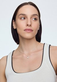 Złoty naszyjnik z okrągłym białym zawieszką noszony na beżowym topie bez rękawów z czarnymi wykończeniami. Gładka faktura, delikatny wzór łańcuszka.