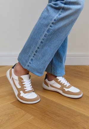 Person wearing white and beige sneakers with white laces and blue jeans, standing on a wooden floor near a white wall.