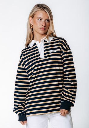 Mujer rubia vistiendo una camiseta de rugby de manga larga a rayas azul marino, blanca y mostaza con cuello blanco y pantalones blancos, mirando hacia la derecha.