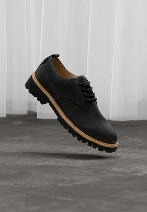 Schwarzer Leder-Schnürschuh mit braunem Rand und klobiger schwarzer Sohle, der über einem grauen Betonboden und weißen transparenten Vorhängen schwebt.