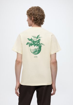 Personne aux cheveux bouclés portant un t-shirt crème avec un motif vert d'un pot de basketball avec des feuilles dans le dos.