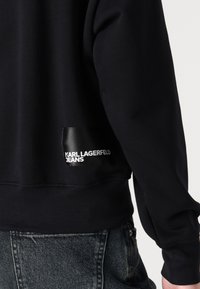 Tamsiai mėlynas džemperis su juodu etiketu, ant kurio užrašas "KARL LAGERFELD JEANS" nugaros pusėje. Pagamintas iš minkštos medžiagos, su įtrauktomis rankovėmis ir juosmeniu.