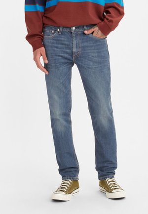 Jean en denim bleu clair avec une coupe slim, zones décolorées, cinq poches et fermeture à bouton. Porté avec des baskets en toile vertes.