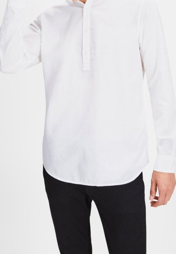 Camicia bianca a maniche lunghe con una pattina bottonata, materiale in cotone morbido, orlo curvo e trama sottile; indossata con pantaloni neri.