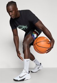 Juodas trumpomis rankovėmis marškinėliai su krepšinio grafika, juodi šortai su vaivorykštės raštu, baltos Nike sportbačiai, laiko oranžinį krepšinį.
