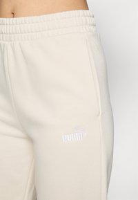 Beige Jogginghose mit elastischem Bund und seitlichen Taschen. Mit einem weißen gestickten Puma-Logo auf dem linken Oberschenkel. Weicher Stoff.