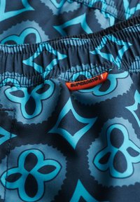 Blauwe zwemshorts met een patroon, voorzien van een elastische tailleband en een rode "SUPERDRY" label. Het materiaal heeft een gladde, glanzende textuur.