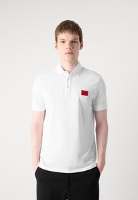 Hvid polo skjorte med krave, korte ærmer og et rødt rektangulært logo patch på venstre bryst. Stoffet ser glat og åndbart ud.