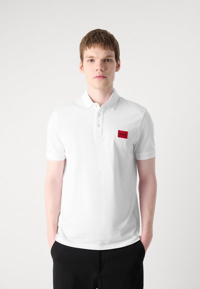 Polo blanc avec un col, des manches courtes et une patch rectangulaire rouge sur la poitrine gauche. Le tissu semble lisse et respirant.