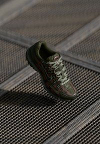 Nike sneaker featuring a mesh design in olive green with red accents, a cushioned sole, and a hexagonal textured pattern throughout.