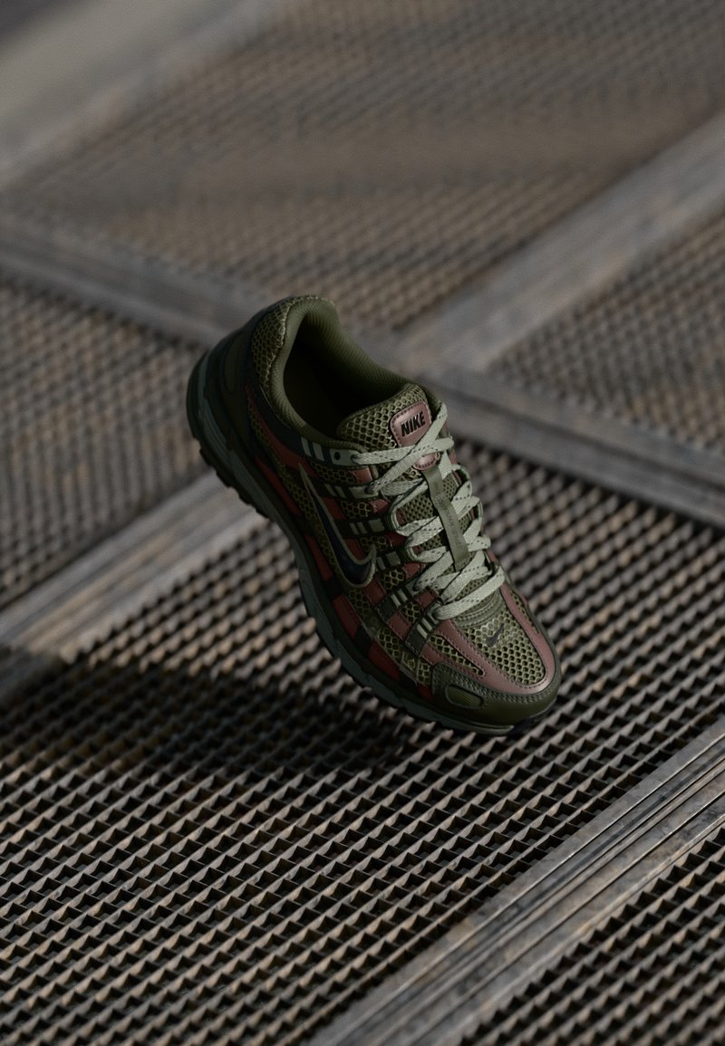 Nike sneaker featuring a mesh design in olive green with red accents, a cushioned sole, and a hexagonal textured pattern throughout.