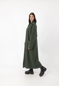 Langer grüner Mantel aus dickem Stoff mit übergroßen Ärmeln, tiefen Taschen und geradem Schnitt, kombiniert mit schwarzen Plateaustiefeln.