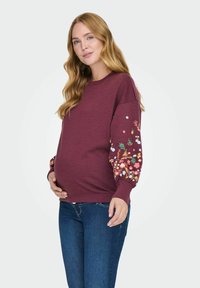 Felpa maternità in cotone bordeaux con ricami floreali sulle maniche. Presenta un collo a giro e polsini a coste. Abbinata a jeans blu.