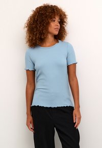 Tricou ribbed de culoare bleu deschis, cu mâneci scurte și tiv ondulat. Materialul pare moale și ajustat, purtat cu pantaloni negri.