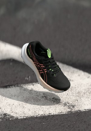 Chaussure de sport noire avec des accents verts, design léger, empeigne texturée et marquage orange vif, sur un fond sombre.