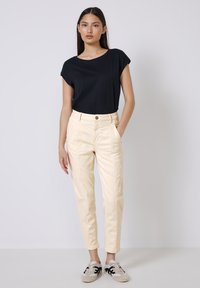 Camiseta negra de manga corta, pantalones de cintura alta en color crema con bolsillos laterales y zapatillas grises con detalles negros, de pie contra un fondo liso.